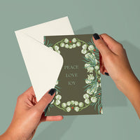 A hand holding a "Peace, Love, Joy" holiday greeting card with white snowdrop flowers on olive green. Illustrated by Lisel Ashlock.