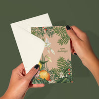 Hands holding an open holiday greeting card with blush background, winter botanicals, citrus fruits, and white bow. Illustrated by Lisel Ashlock.