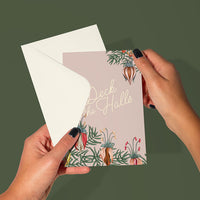 Person holding a "Deck the Halls" holiday greeting card with floral and botanical illustrations against a green background. Illustrated by Lisel Ashlock.