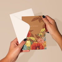 Hands holding open a rust-colored floral birthday greeting card with yellow, red, and pink flowers. Illustrated by Lisel Ashlock.