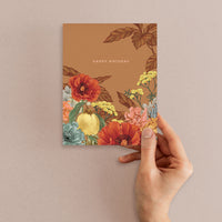 Hand holding a rust-colored Happy Birthday greeting card featuring red poppies, yellow flowers, and botanical illustrations. Illustrated by Lisel Ashlock.