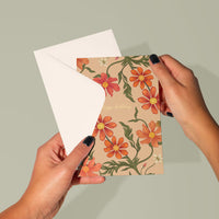 A hand holding open a floral birthday greeting card with red and orange cosmos flowers on a tan background. Illustrated by Lisel Ashlock.