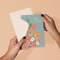 Person holding a teal greeting card with colorful poppy flowers and "happy birthday" text. Illustrated by Lisel Ashlock.
