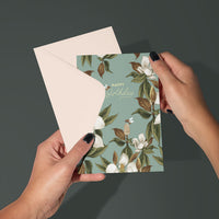 A hand holding a teal birthday greeting card with white magnolia flowers and gold leaves, with matching cream envelope. Illustrated by Lisel Ashlock.