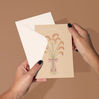 A hand holding an open greeting card with a beige background featuring a pink and orange flower bouquet illustration and "celebrating you" text. Illustrated by Lisel Ashlock.