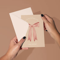 Hand holding a beige birthday greeting card with pink bow illustration and "celebrating you happy birthday" text. Illustrated by Lisel Ashlock.