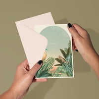 Person holding open a "Thank You" greeting card featuring tropical foliage and a blue circle. Illustrated by Lisel Ashlock.