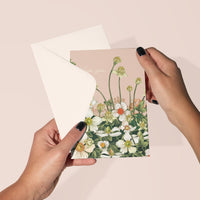 Hands holding open a pink greeting card with "love you" text and white anemone flowers illustrated by Lisel Ashlock.