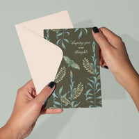 Hands holding an open sympathy greeting card with botanical design and "keeping you in my thoughts" text. Illustrated by Lisel Ashlock.