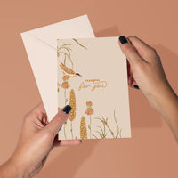 Person holding a "Thankful for you" greeting card with illustrated wildflowers and a bird against a tan background. Illustrated by Lisel Ashlock.