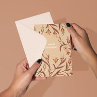 Hand holding a peach-toned birthday greeting card with brown floral illustrations and white envelope against tan background. Illustrated by Lisel Ashlock.