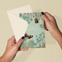Two hands holding an open greeting card with botanical illustration and orange butterflies, text reads "TO THE Happy Couple". Illustrated by Lisel Ashlock.