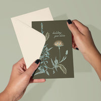 Hands holding olive green greeting card with botanical illustrations and text "holding you close" against cream envelope. Illustrated by Lisel Ashlock.