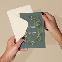 Hands holding an open greeting card with "Always and Forever" text and botanical wreath design on sage green. Illustrated by Lisel Ashlock.