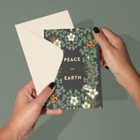 Hands holding a "Peace on Earth" greeting card with botanical floral design on dark background. Illustrated by Lisel Ashlock.