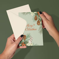 Hands holding a mint green Merry Christmas greeting card with pine cones, holly, berries, and a hare illustration by Lisel Ashlock.