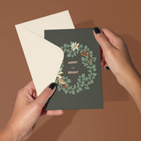 Hands holding a dark green greeting card with "Merry and Bright" text surrounded by a festive wreath illustration. Illustrated by Lisel Ashlock.