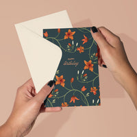 A woman's hands holding an open greeting card with orange flowers on dark background saying "Happy birthday" Illustrated by Lisel Ashlock.