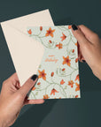 Hands holding a Happy Birthday greeting card with orange flowers and green vines on sea foam background. Illustrated by Lisel Ashlock.