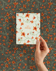 Hand holding a birthday greeting card with orange flowers on cream background against matching patterned wallpaper. Illustrated by Lisel Ashlock.