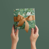 Two hands holding botanical gift wrap featuring green foliage and white flowers tied with a tan ribbon bow. Illustrated by Lisel Ashlock.