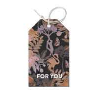 A gift tag featuring an illustrated botanical and wildlife pattern with flowers, leaves, monkeys, and butterflies on a dark background with "FOR YOU" text. Illustrated by Lisel Ashlock.