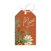 A rust-colored gift tag decorated with pinecones, evergreen branches, white flowers, and orange berries with "For you" text. Illustrated by Lisel Ashlock.