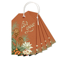 A fan-shaped stack of rust-colored gift tags with "For you" text and botanical illustrations including pinecones and flowers, tied with white string. Illustrated by Lisel Ashlock.