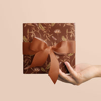 Hand holding a square wrapped gift with rust-colored botanical pattern and coordinating ribbon bow. Illustrated by Lisel Ashlock.
