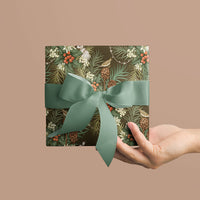 A hand holding a square gift wrapped in Holly & Hare moss patterned paper with a sage green ribbon bow. Illustrated by Lisel Ashlock.