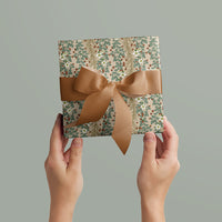 Two hands holding a wrapped gift with "Boughs & Berries in Blush" wrapping paper and tan ribbon bow. Illustrated by Lisel Ashlock.
