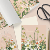 Flat lay of pink anemone floral gift wrap, cards, envelope, scissors, tape, and pen on beige background. Illustrated by Lisel Ashlock.