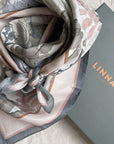 Folded Angelite silk cotton scarf in cream, gray, and mauve tones with botanical leaf motifs beside LINNA packaging. Illustrated by Lisel Ashlock.