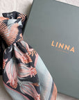 A folded silk cotton scarf with floral botanical print in rust, pink, and cream tones next to its gray LINNA California box. Illustrated by Lisel Ashlock.