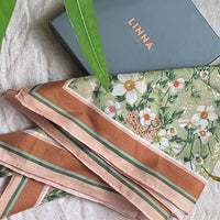 Folded silk cotton scarves in pink and brown tones with green striped accents and floral anemone pattern, displayed with LINNA packaging. Illustrated by Lisel Ashlock.