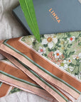 Folded silk cotton scarves in pink and brown tones with green striped accents and floral anemone pattern, displayed with LINNA packaging. Illustrated by Lisel Ashlock.