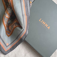 A folded apricot filigree silk cotton scarf next to its Linna California branded box on a neutral surface.