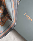 A folded apricot filigree silk cotton scarf next to its Linna California branded box on a neutral surface.