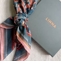 Patterned silk cotton scarf in blue with red roses tied around a wrist next to gray LINNA box. Illustrated by Lisel Ashlock.