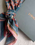 Patterned silk cotton scarf in blue with red roses tied around a wrist next to gray LINNA box. Illustrated by Lisel Ashlock.