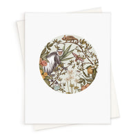 A circular illustration of a guereza monkey surrounded by botanical elements including flowers, fruits, and foliage with a chameleon perched above, on a blank greeting card. Illustrated by Lisel Ashlock.