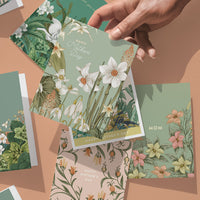 Hand holding a green greeting card with white daffodils and "Happy Father's Day" text among coordinating floral cards. Illustrated by Lisel Ashlock.