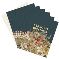 A fanned stack of dark green greeting cards with gold "Season's Greetings" text and botanical illustrations. Illustrated by Lisel Ashlock.