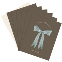A stack of brown greeting cards with a blue bow illustration and "celebrating you happy birthday" text. Illustrated by Lisel Ashlock.