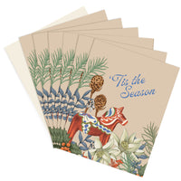 Stack of holiday greeting cards featuring a red Dala horse holding pine cones surrounded by botanical illustrations and "'Tis the Season" text. Illustrated by Lisel Ashlock.