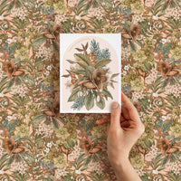 A hand holding a Season's Greetings card featuring botanical hellebore flowers against patterned background. Illustrated by Lisel Ashlock.