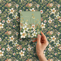 A hand holding a thank you greeting card with white flowers on sage green background against a matching floral patterned surface. Illustrated by Lisel Ashlock.