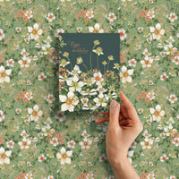 Hand holding a green birthday greeting card with white flowers and gold text against a green floral background. Illustrated by Lisel Ashlock.