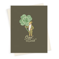 A leprechaun in brown clothing holding a large four-leaf clover with "Good Luck" text on a greeting card. Illustrated by Lisel Ashlock.