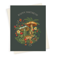 A birthday greeting card featuring illustrated mushrooms, a hare, ferns, raspberries, and wildflowers on a dark green background. Illustrated by Lisel Ashlock.